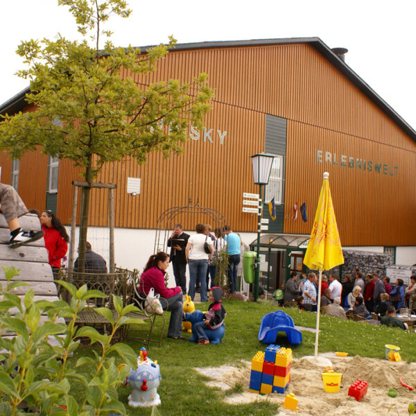An einem bewölkten Tag versammeln sich Menschen vor einem Holzgebäude in der Nähe eines Spielplatzes mit Spielzeug und Sand, umgeben von Grün und gelben Regenschirmen.