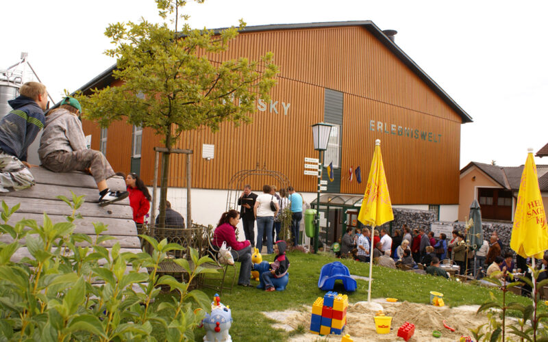 An einem bewölkten Tag versammeln sich Menschen vor einem Holzgebäude in der Nähe eines Spielplatzes mit Spielzeug und Sand, umgeben von Grün und gelben Regenschirmen.