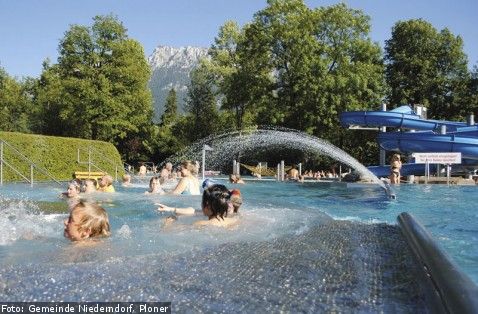Außenschwimmbad mit Menschen, die das Wasser genießen, einer Wasserrutsche und einer blauen Wasserrutsche, umgeben von Bäumen und Bergen im Hintergrund.