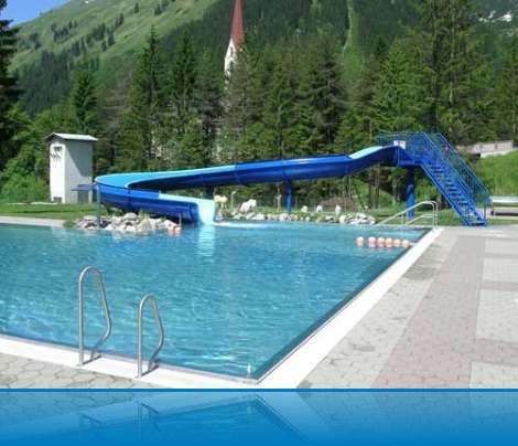 Außenschwimmbad mit Metallleiter und blauer Rutsche, umgeben von Bäumen und Bergen; im Hintergrund ist ein Kirchturm zu sehen.