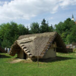 Eine kleine, strohgedeckte Hütte steht auf einer Wiese mit Bäumen und einem Kirchturm im Hintergrund unter einem teilweise bewölkten Himmel.
