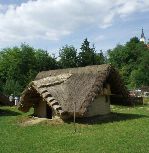 Eine kleine, strohgedeckte Hütte steht auf einer Wiese mit Bäumen und einem Kirchturm im Hintergrund unter einem teilweise bewölkten Himmel.