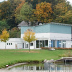 Modernes weiß-blaues Gebäude mit großen Fenstern, umgeben von Bäumen mit Herbstlaub, neben einer Wiese und einem Holzsteg am Wasser.