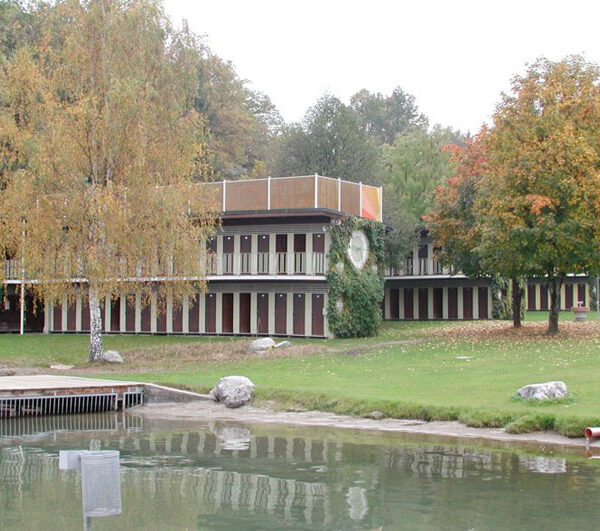 Ein modernes, zweistöckiges Gebäude mit vielen vertikalen Fenstern steht an einem Teich, umgeben von grünem Gras, Bäumen und etwas Herbstlaub.