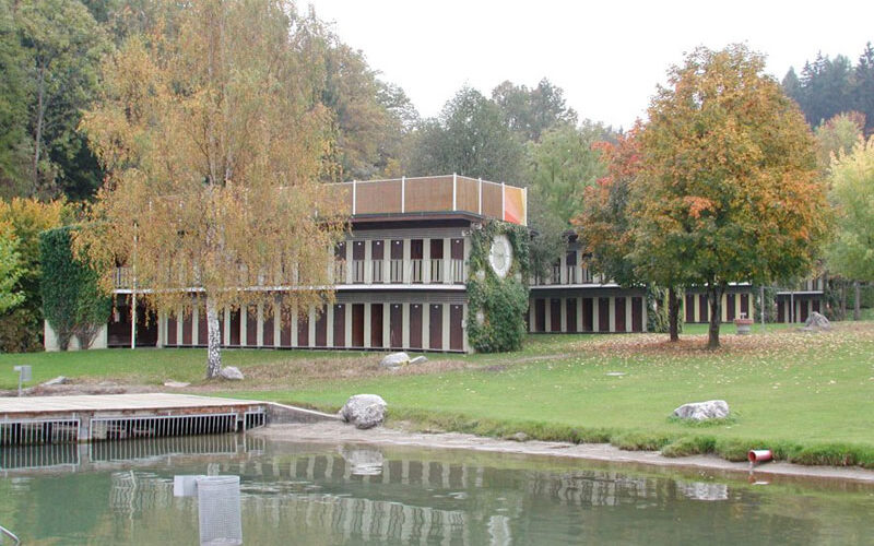 Ein modernes, zweistöckiges Gebäude mit vielen vertikalen Fenstern steht an einem Teich, umgeben von grünem Gras, Bäumen und etwas Herbstlaub.