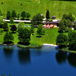 Ein Park am Seeufer mit grünen Rasenflächen, vereinzelten Bäumen, einem Steg, einem kleinen Sandstrand, geparkten Autos und Menschen, die sich an einem sonnigen Tag im Freien entspannen.
