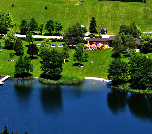 Ein Park am Seeufer mit grünen Rasenflächen, vereinzelten Bäumen, einem Steg, einem kleinen Sandstrand, geparkten Autos und Menschen, die sich an einem sonnigen Tag im Freien entspannen.
