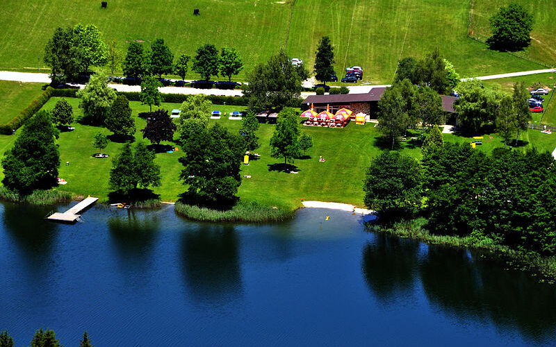 Ein Park am Seeufer mit grünen Rasenflächen, vereinzelten Bäumen, einem Steg, einem kleinen Sandstrand, geparkten Autos und Menschen, die sich an einem sonnigen Tag im Freien entspannen.