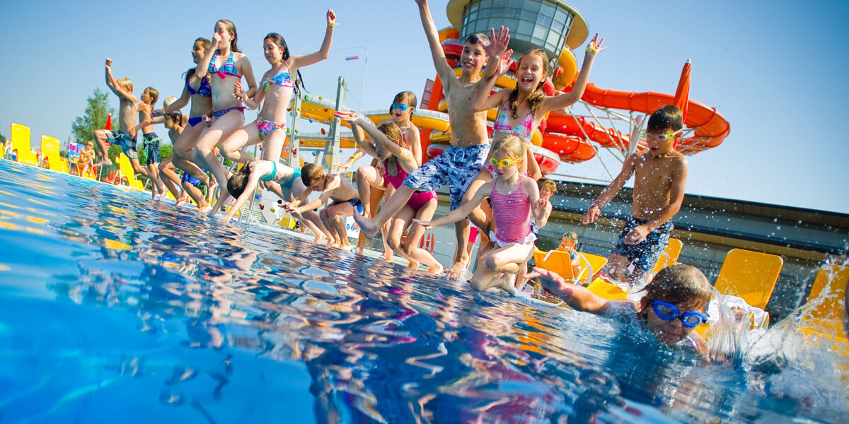Eine Gruppe von Kindern in Badeanzügen springt in ein Freibad, im Hintergrund sind Wasserrutschen und Liegestühle zu sehen.