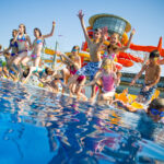 Eine Gruppe von Kindern in Badeanzügen springt in ein Freibad, im Hintergrund sind Wasserrutschen und Liegestühle zu sehen.