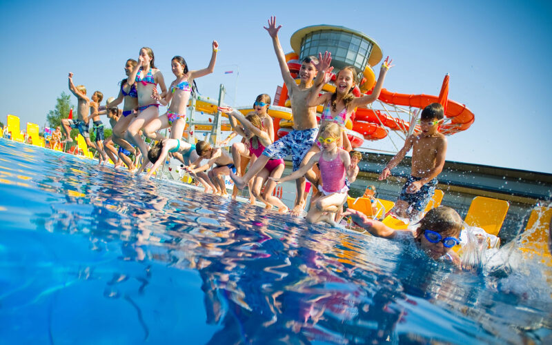 Eine Gruppe von Kindern in Badeanzügen springt in ein Freibad, im Hintergrund sind Wasserrutschen und Liegestühle zu sehen.