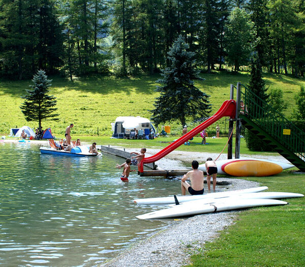 Menschen genießen einen Freizeitbereich am See mit einer roten Rutsche, Tretbooten, Kanus und Kajaks; im Hintergrund sind Zelte und Bäume zu sehen.
