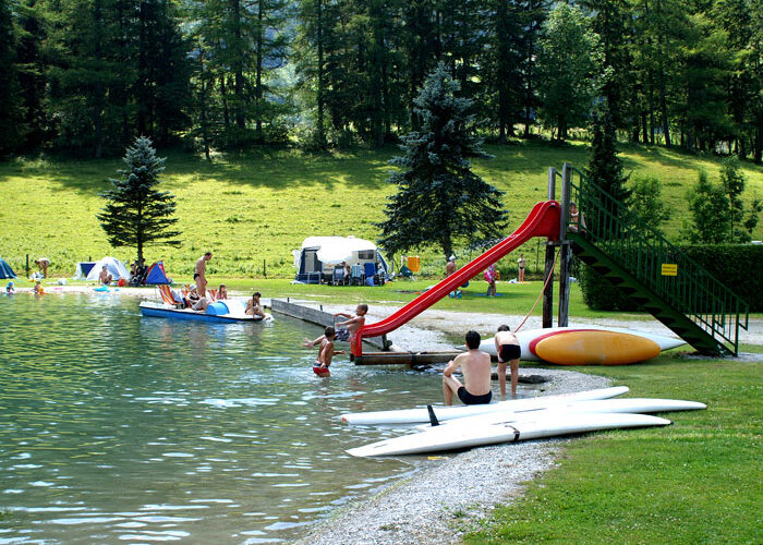 Menschen genießen einen Freizeitbereich am See mit einer roten Rutsche, Tretbooten, Kanus und Kajaks; im Hintergrund sind Zelte und Bäume zu sehen.