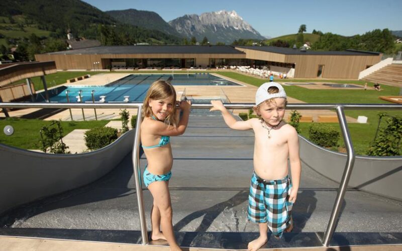 Zwei kleine Kinder in Badekleidung stehen an der Spitze einer Wasserrutsche in einem Freibadkomplex mit Bergen im Hintergrund.