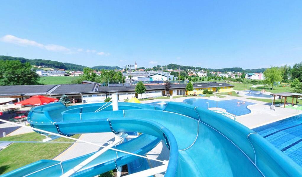 Eine große blaue Wasserrutsche führt zu einem Außenpool mit schattigen Sitzgelegenheiten, umgeben von grüner Landschaft und Gebäuden unter einem klaren Himmel.