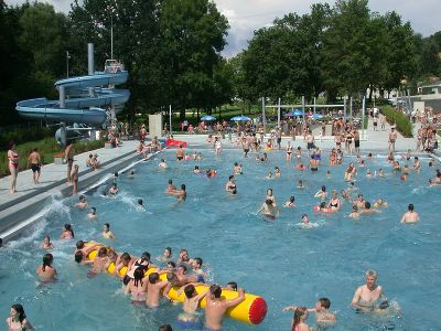 Überfülltes Freibad mit vielen Menschen im Wasser und auf der Terrasse, einem großen aufblasbaren Schwimmkörper und einer blauen Wasserrutsche im Hintergrund.