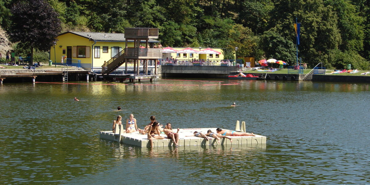 Menschen entspannen sich auf einer schwimmenden Plattform in einem See mit einem Gebäude und Bäumen im Hintergrund an einem sonnigen Tag.