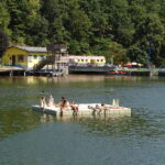 Menschen entspannen sich auf einer schwimmenden Plattform in einem See mit einem Gebäude und Bäumen im Hintergrund an einem sonnigen Tag.