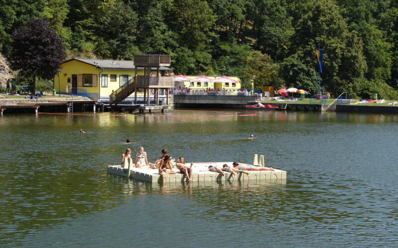 Menschen entspannen sich auf einer schwimmenden Plattform in einem See mit einem Gebäude und Bäumen im Hintergrund an einem sonnigen Tag.