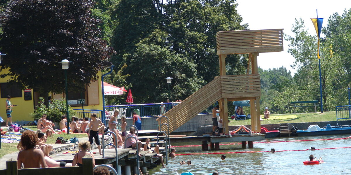 An einem See mit Holzstegen und einem Sprungturm kann man sich an sonnigen Tagen entspannen und schwimmen, umgeben von Bäumen und Grasflächen.