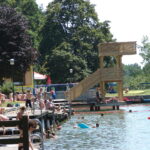 An einem See mit Holzstegen und einem Sprungturm kann man sich an sonnigen Tagen entspannen und schwimmen, umgeben von Bäumen und Grasflächen.