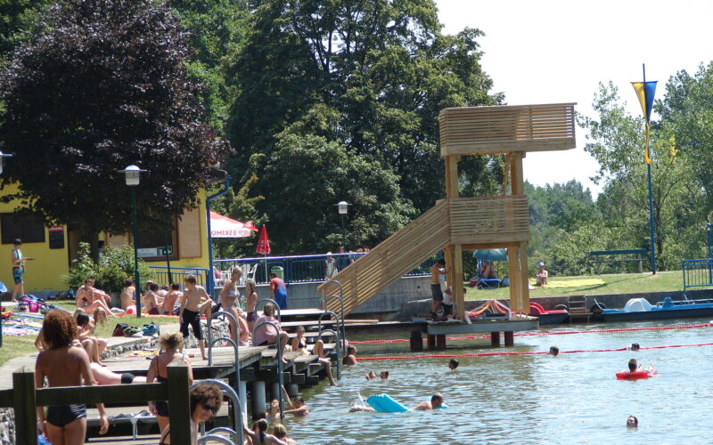 An einem See mit Holzstegen und einem Sprungturm kann man sich an sonnigen Tagen entspannen und schwimmen, umgeben von Bäumen und Grasflächen.
