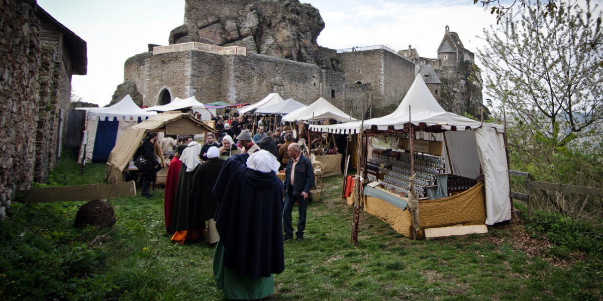 Menschen besuchen einen mittelalterlichen Jahrmarkt mit Zelten und Marktständen in der Nähe einer historischen Steinburg auf einer Wiese.