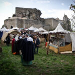 Menschen besuchen einen mittelalterlichen Jahrmarkt mit Zelten und Marktständen in der Nähe einer historischen Steinburg auf einer Wiese.