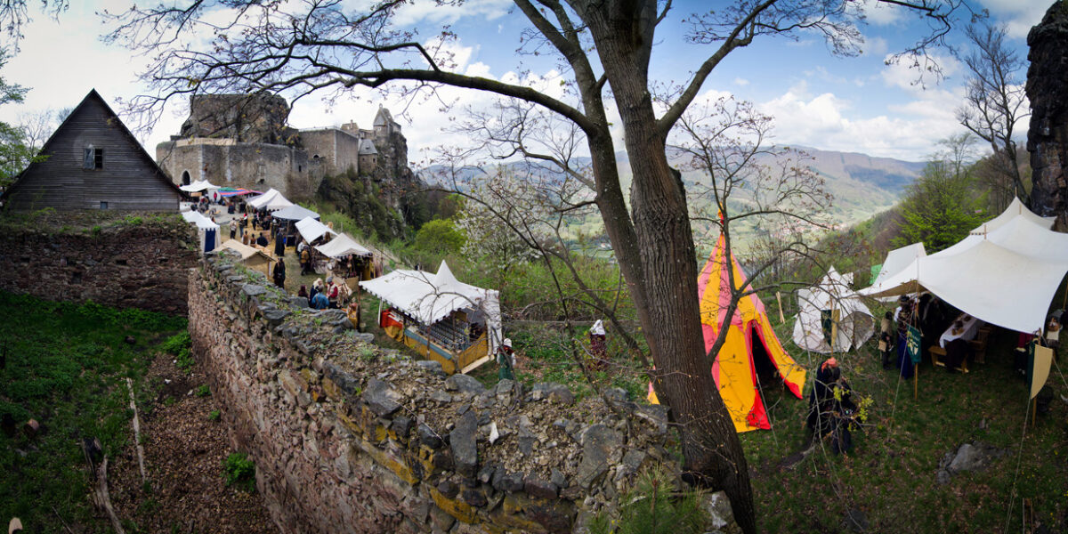 Ein mittelalterlicher Markt mit Zelten und Menschen ist entlang der Steinmauern einer alten Burg auf einem Hügel aufgebaut, umgeben von Bäumen und fernen Bergen.