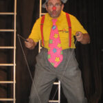 Eine als Clown verkleidete Person mit einem bunten Hut, einer großen geblümten Krawatte, einem gelben Hemd und einer karierten Hose führt auf der Bühne eine Diabolo-Nummer vor.