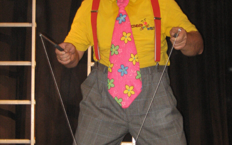 Eine als Clown verkleidete Person mit einem bunten Hut, einer großen geblümten Krawatte, einem gelben Hemd und einer karierten Hose führt auf der Bühne eine Diabolo-Nummer vor.