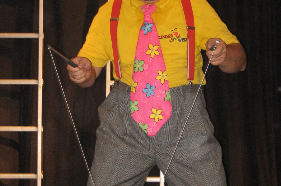 Eine als Clown verkleidete Person mit einem bunten Hut, einer großen geblümten Krawatte, einem gelben Hemd und einer karierten Hose führt auf der Bühne eine Diabolo-Nummer vor.