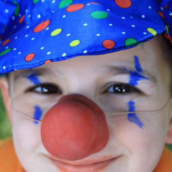 Kind mit blau gepunktetem Clownshut, roter Clownsnase, orangefarbenem Hemd und blauer Gesichtsbemalung, das in die Kamera lächelt.