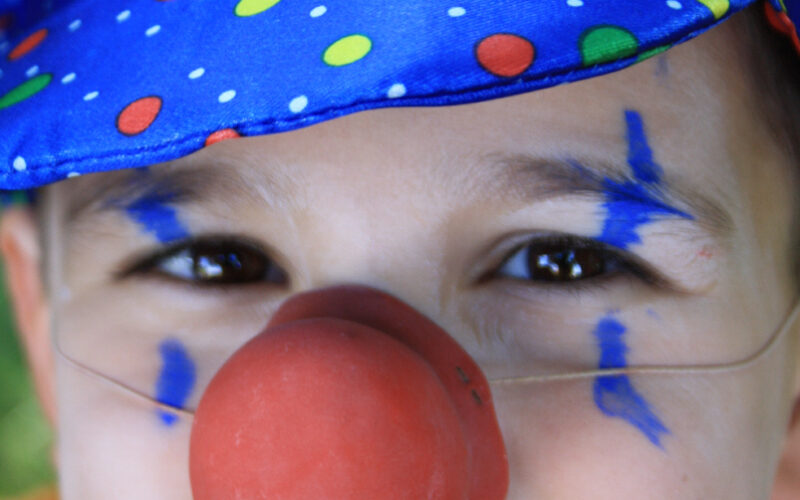 Kind mit blau gepunktetem Clownshut, roter Clownsnase, orangefarbenem Hemd und blauer Gesichtsbemalung, das in die Kamera lächelt.