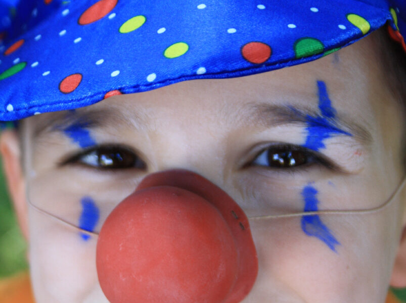 Kind mit blau gepunktetem Clownshut, roter Clownsnase, orangefarbenem Hemd und blauer Gesichtsbemalung, das in die Kamera lächelt.