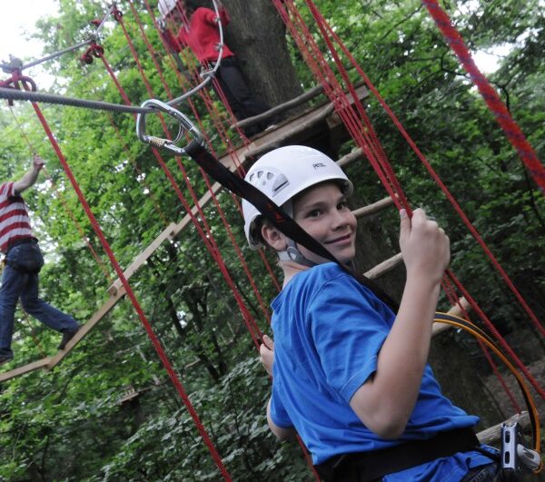 Kinder, die Helme und Klettergurte tragen, nehmen an einem Hochseilgarten im Freien teil und klettern und balancieren auf erhöhten Plattformen zwischen Bäumen.