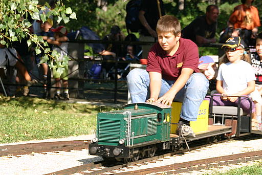 Ein Junge sitzt auf einem kleinen grünen Aufsitzzug und fährt ihn, wobei er mehrere Kinder auf einer Miniatureisenbahn im Freien hinter sich herzieht.