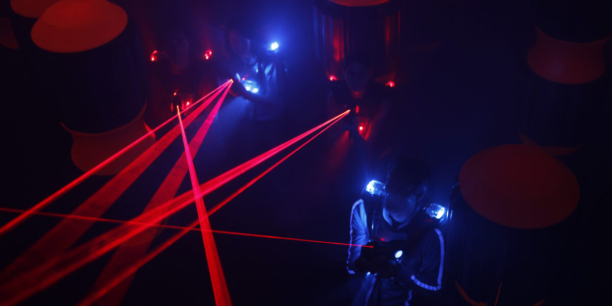 Menschen spielen in einem dunklen Raum Laser-Tag, sie tragen Westen mit leuchtenden blauen und roten Lichtern, während rote Laserstrahlen die Szene durchkreuzen.