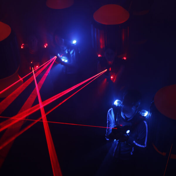 Menschen spielen in einem dunklen Raum Laser-Tag, sie tragen Westen mit leuchtenden blauen und roten Lichtern, während rote Laserstrahlen die Szene durchkreuzen.