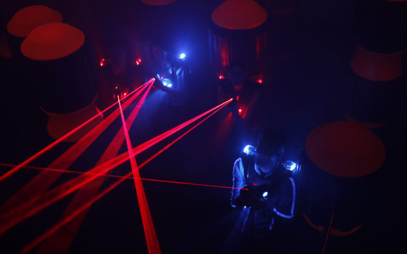 Menschen spielen in einem dunklen Raum Laser-Tag, sie tragen Westen mit leuchtenden blauen und roten Lichtern, während rote Laserstrahlen die Szene durchkreuzen.