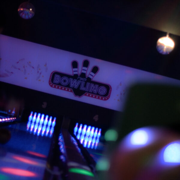 Eine leuchtende Bowlingbahn mit Neonlicht und einem Schild mit der Aufschrift "Bowling" über den Bahnen; am Ende der Bahnen sind Bowlingpins beleuchtet.