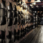 An den Holzwänden entlang eines schwach beleuchteten Korridors mit Holzboden sind Reihen von schwarzen Metallpanzern und Helmen angebracht.