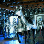 Ein Raum voller mittelalterlicher Rüstungen, in dessen Mitte ein vollständig gepanzertes Pferd zu sehen ist, während im Hintergrund weitere Rüstungen die Wände säumen.