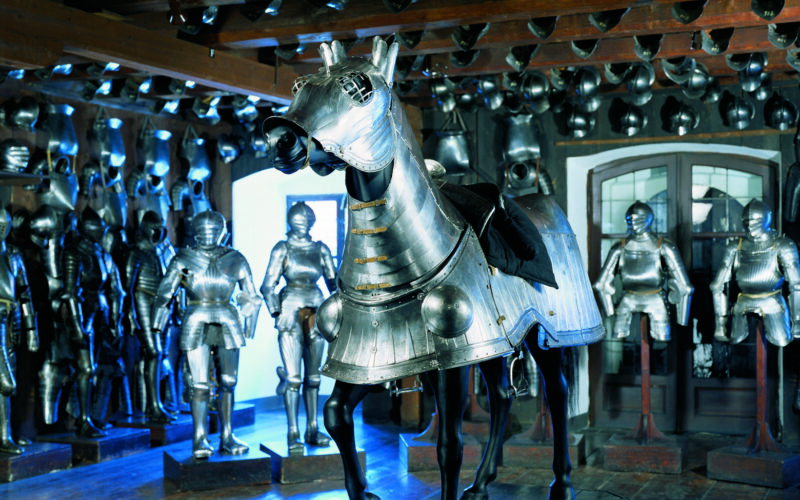 Ein Raum voller mittelalterlicher Rüstungen, in dessen Mitte ein vollständig gepanzertes Pferd zu sehen ist, während im Hintergrund weitere Rüstungen die Wände säumen.