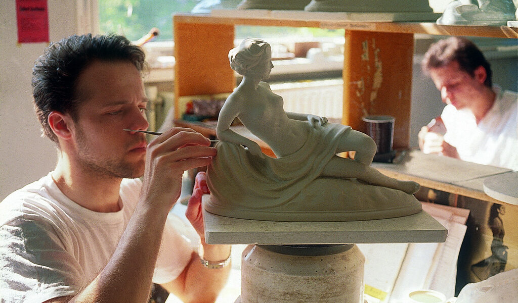 Ein Mann formt sorgfältig Details an einer liegenden weiblichen Statue in einer Werkstatt, während eine andere Person im Hintergrund arbeitet.
