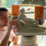 Ein Mann formt sorgfältig Details an einer liegenden weiblichen Statue in einer Werkstatt, während eine andere Person im Hintergrund arbeitet.