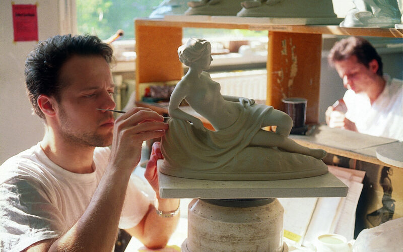 Ein Mann formt sorgfältig Details an einer liegenden weiblichen Statue in einer Werkstatt, während eine andere Person im Hintergrund arbeitet.