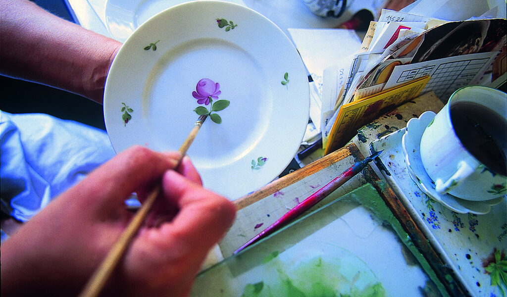 Eine Person malt mit einem dünnen Pinsel eine violette Blume mit grünen Blättern auf einen weißen Teller, in der Nähe befinden sich Malutensilien und eine Tasse Tee.