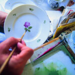 Eine Person malt mit einem dünnen Pinsel eine violette Blume mit grünen Blättern auf einen weißen Teller, in der Nähe befinden sich Malutensilien und eine Tasse Tee.