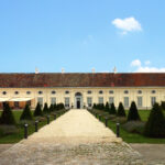 Ein langgestrecktes, einstöckiges Gebäude mit einem Ziegeldach und mehreren Fenstern steht am Ende eines Kiesweges, der von beschnittenen Büschen und Lampen gesäumt ist, unter einem klaren blauen Himmel.
